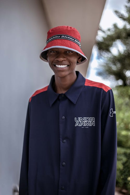 Positive African American young male in hat standing near white wall and looking at camera with toothy smile