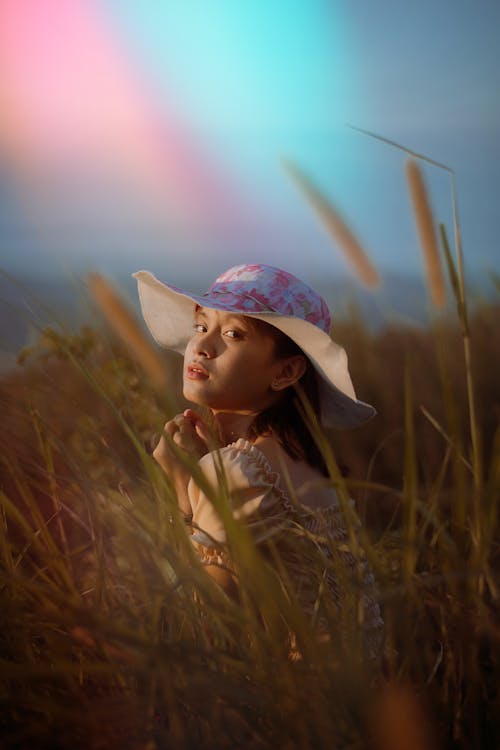 Teen girl in casual clothes and hat sitting in grassy field and enjoying nature in summer day while looking at camera