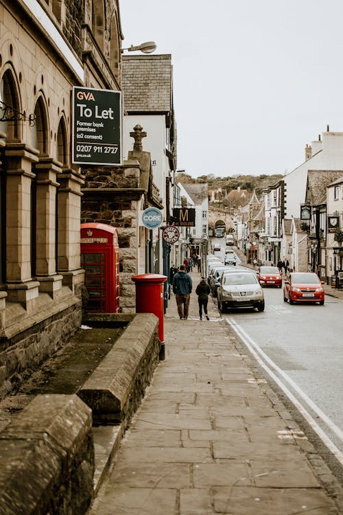 Free Full length people walking trough pathway past road with parked and moving cars and vintage buildings with restaurants emblems and red public call box on English street in daytime Stock Photo