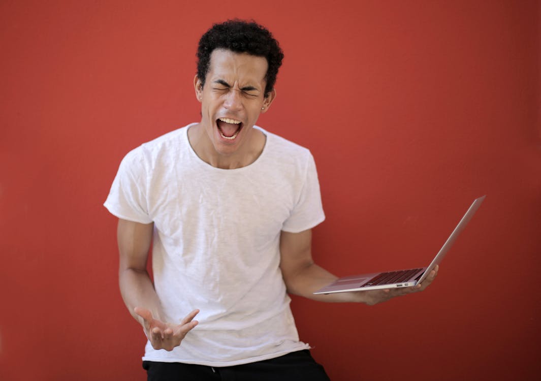 Free Guy with opened mouth and closed eyes screaming madly while standing with laptop against red background Stock Photo