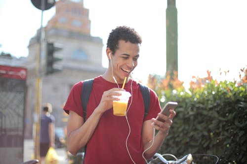 Free Happy ethnic guy in casual wear and with backpack having cup of fresh juice using smartphone while standing on street and listening to music Stock Photo