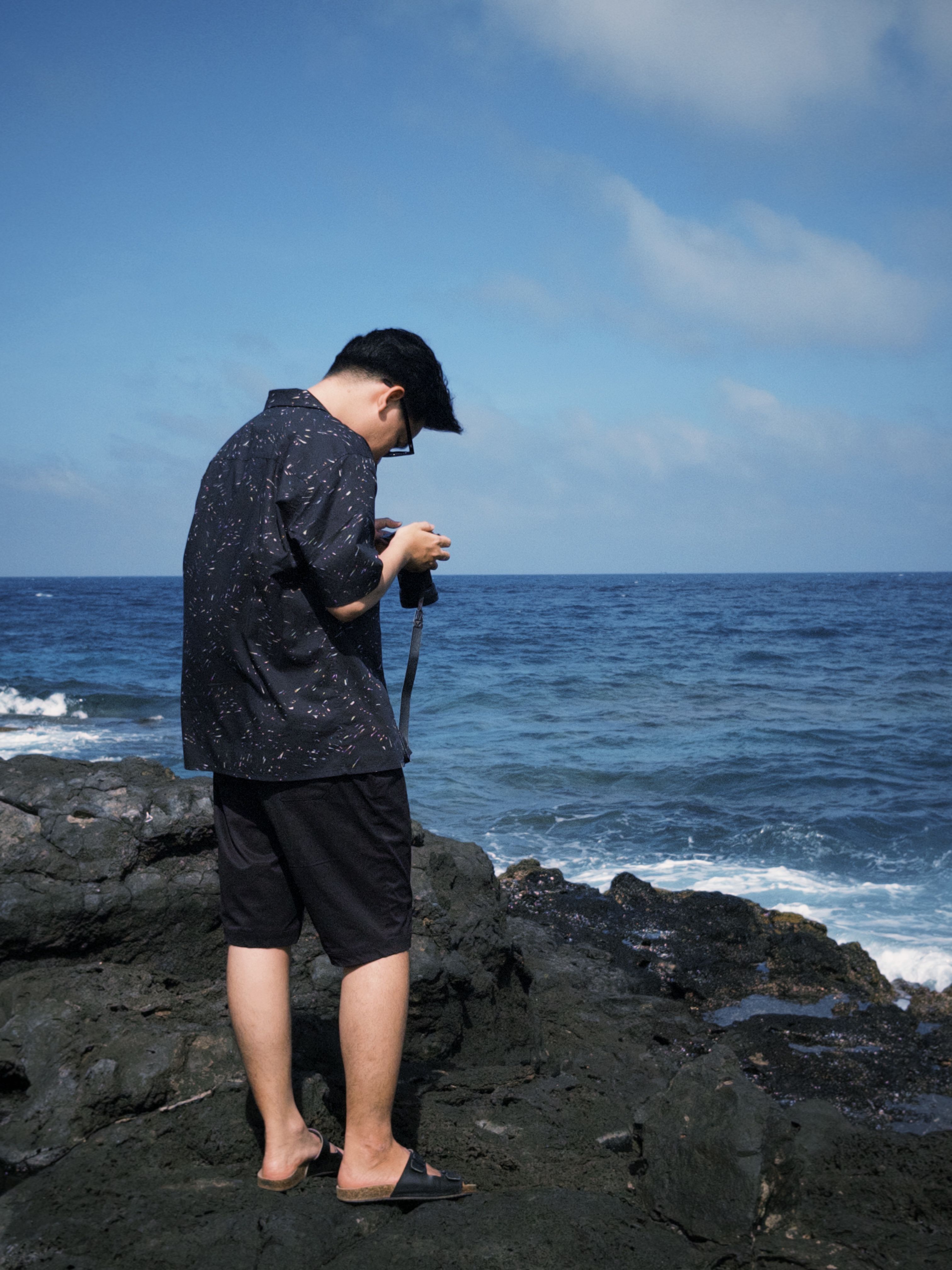 A man takes pictures of the ocean from a rocky coastline under a bright blue sky.