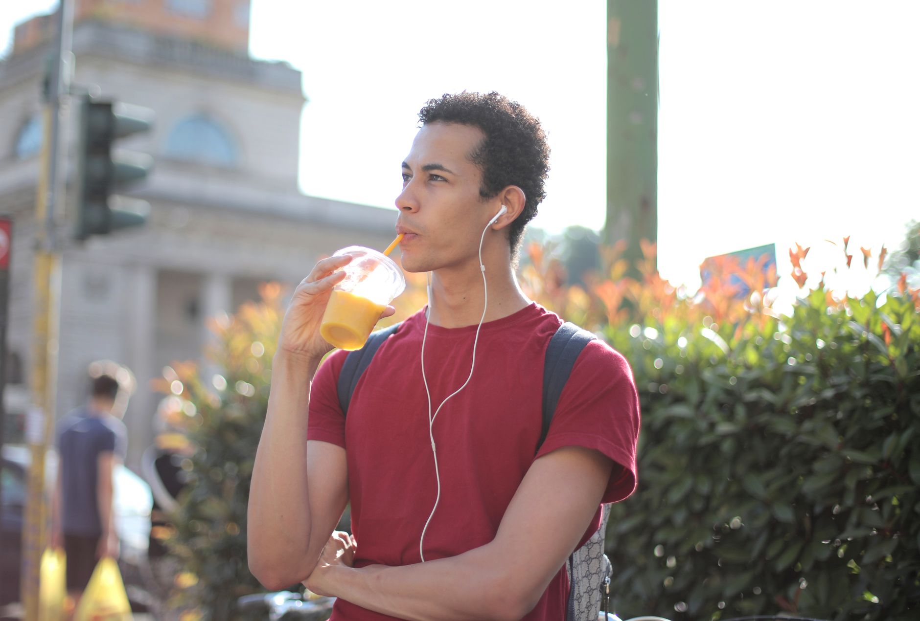 Young man in red shirt sipping a smoothie while listening to music outdoors.