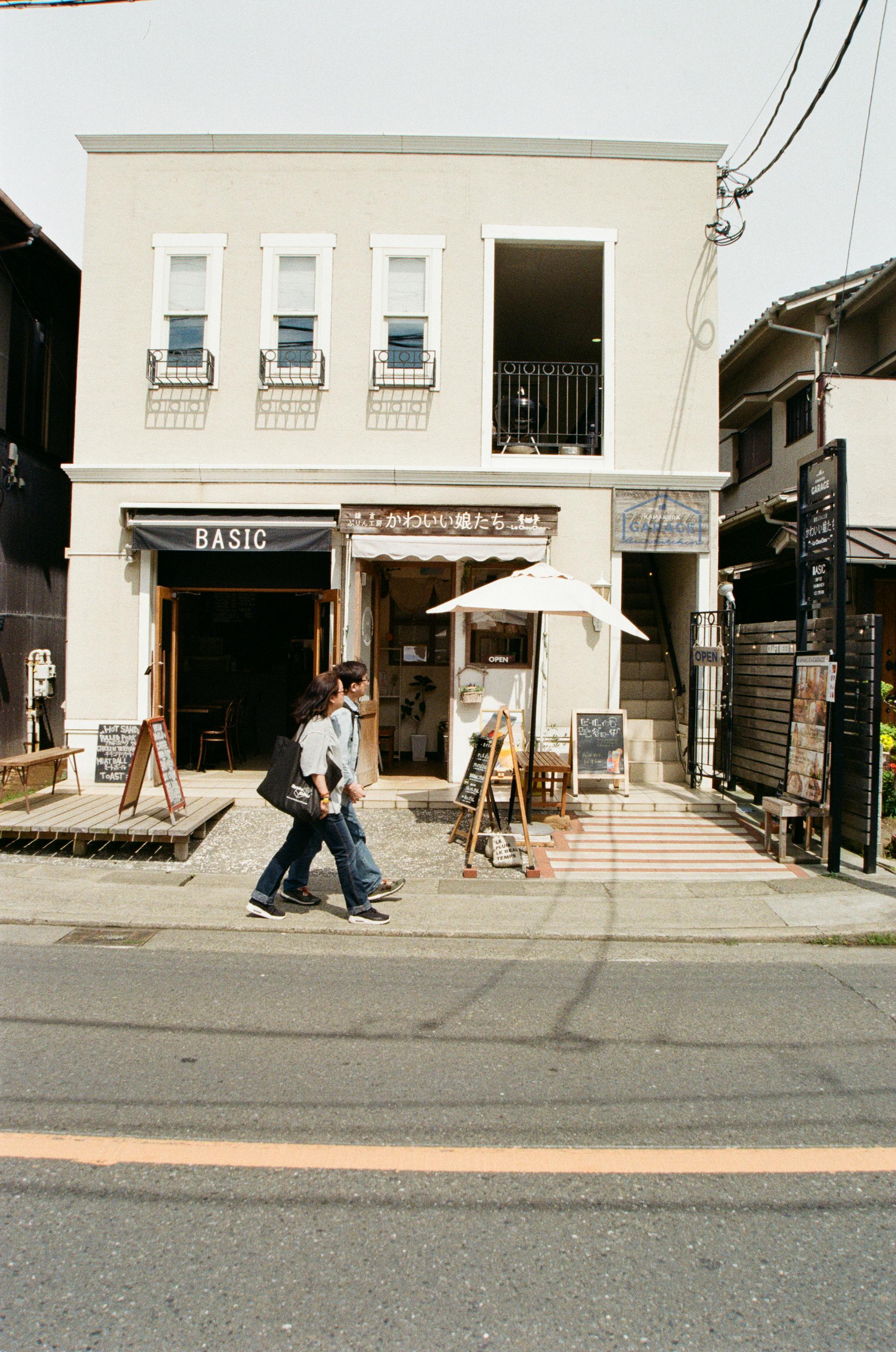 A couple holding hands while walking past quaint stores on a sunny street in Kamakura, Japan.