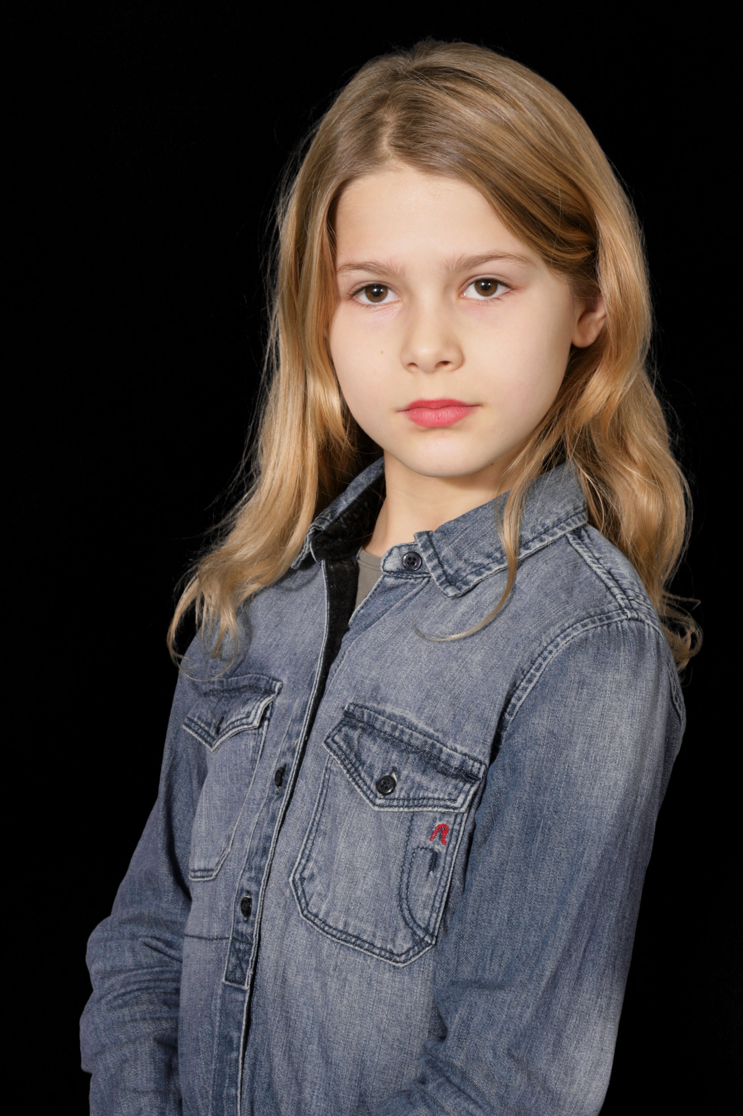 Portrait of a young girl with blond hair wearing a denim shirt against a black background.