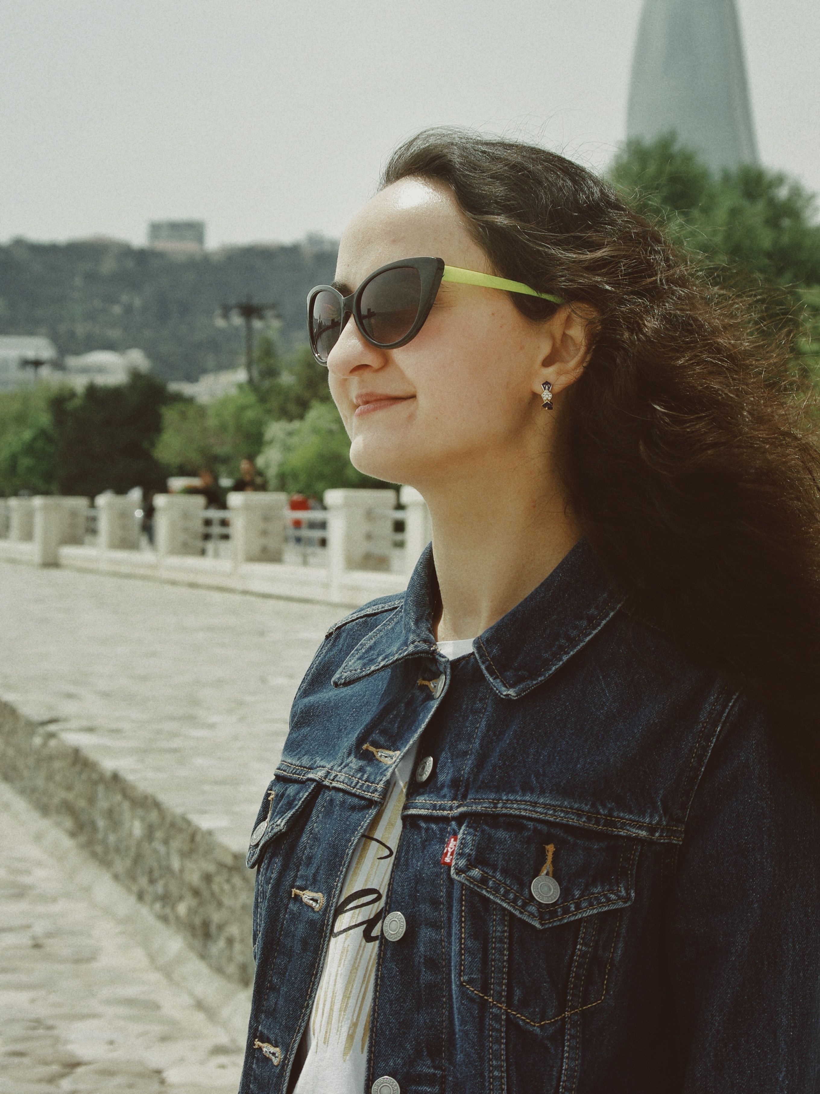 Portrait of a woman wearing sunglasses and denim jacket in Baku with scenic backdrop.