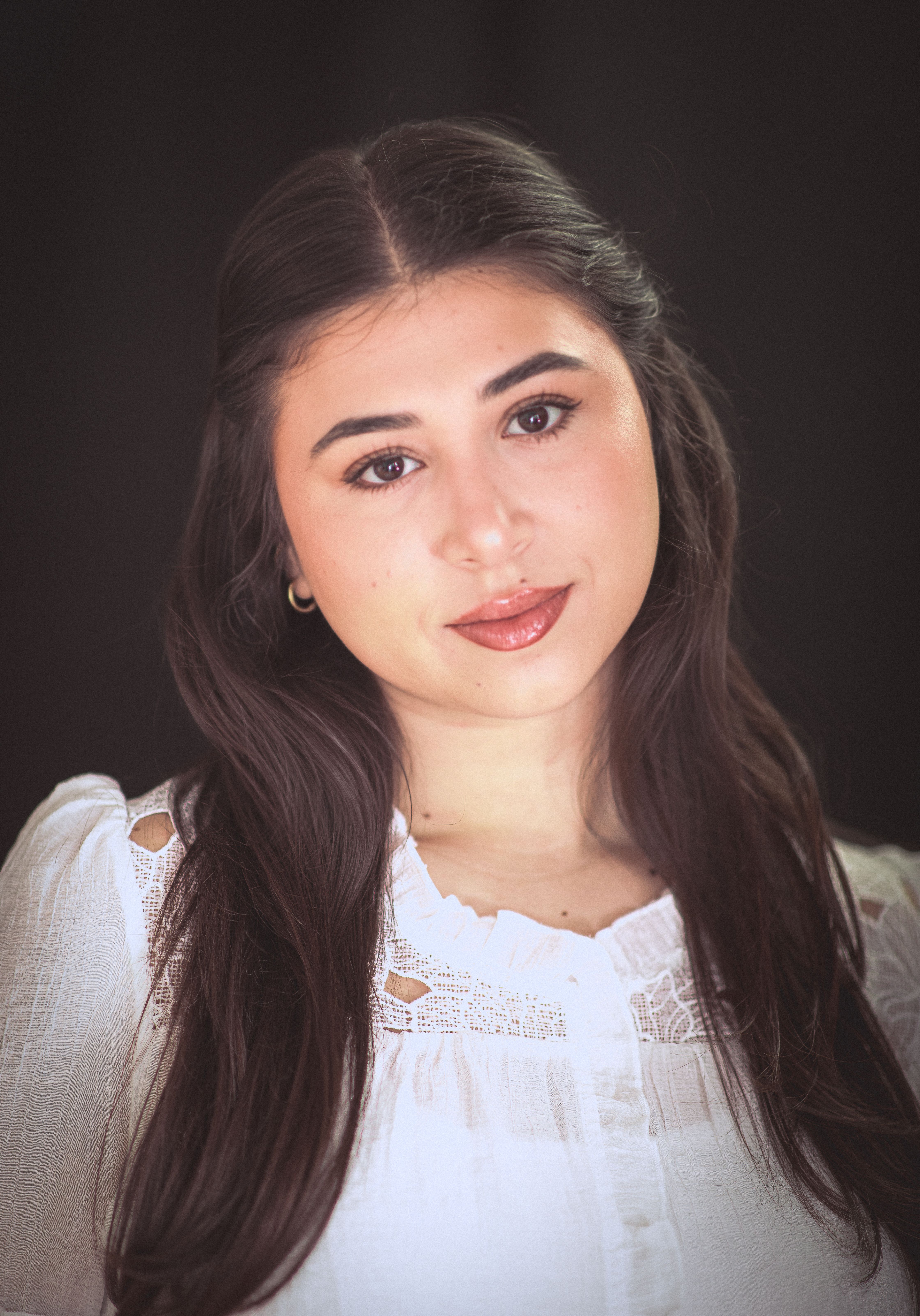 Close-up portrait of a young woman with long hair, wearing a white blouse, against a dark background.