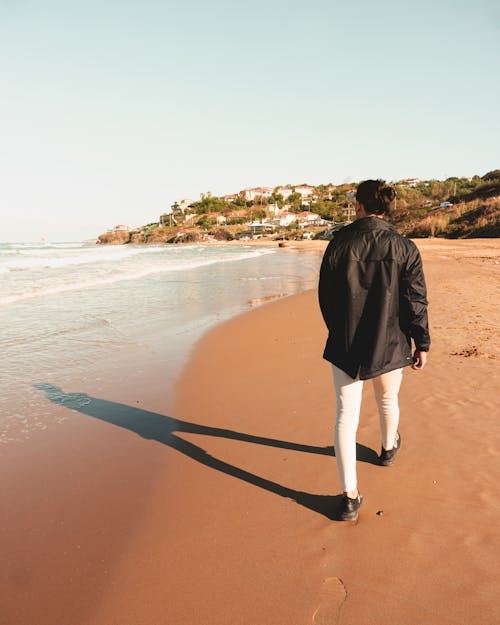 Man in Black Jacket and White Pants Walking on Beach