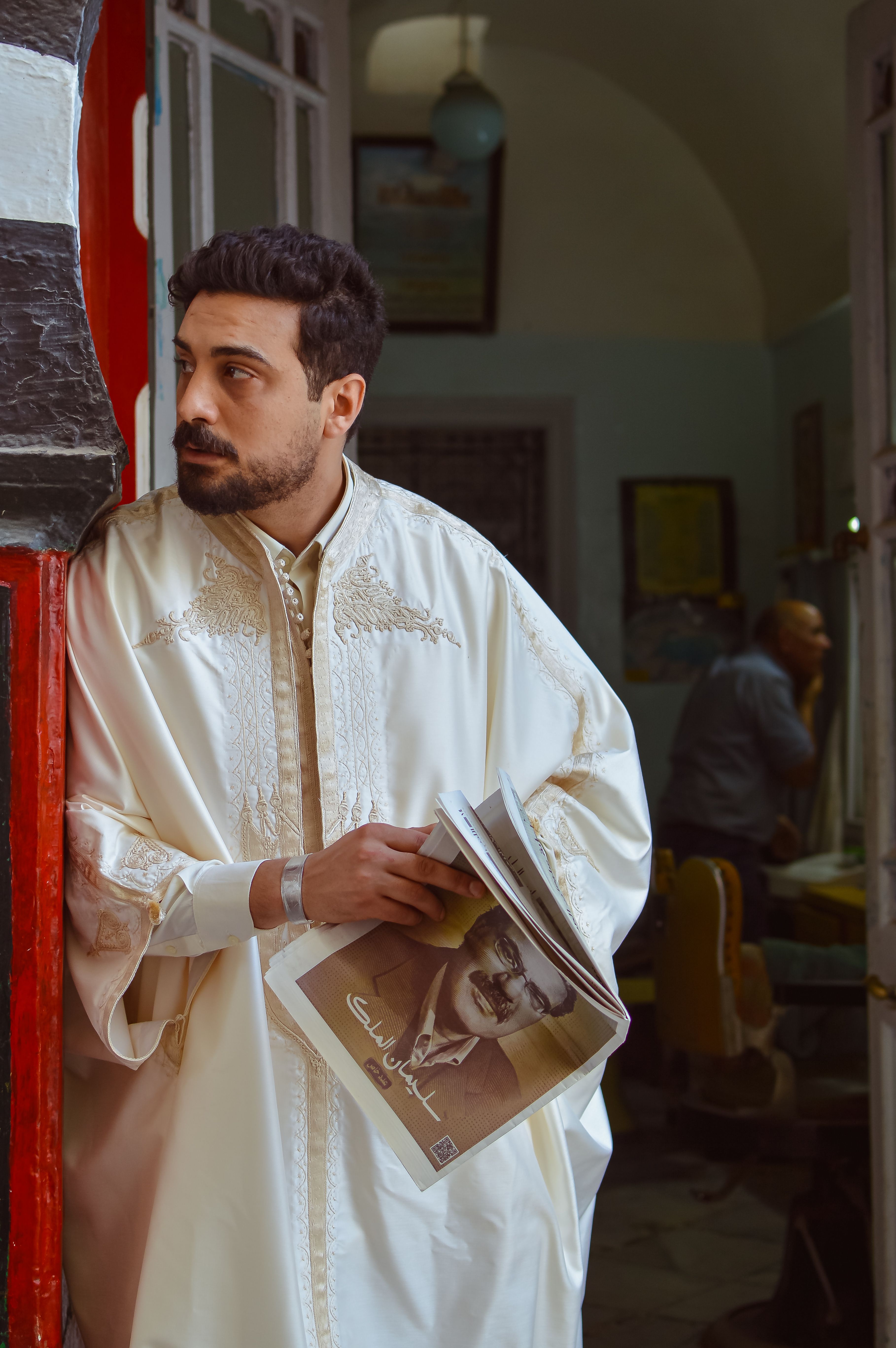 Man in traditional attire holds a newspaper, standing thoughtfully in a doorway indoors.