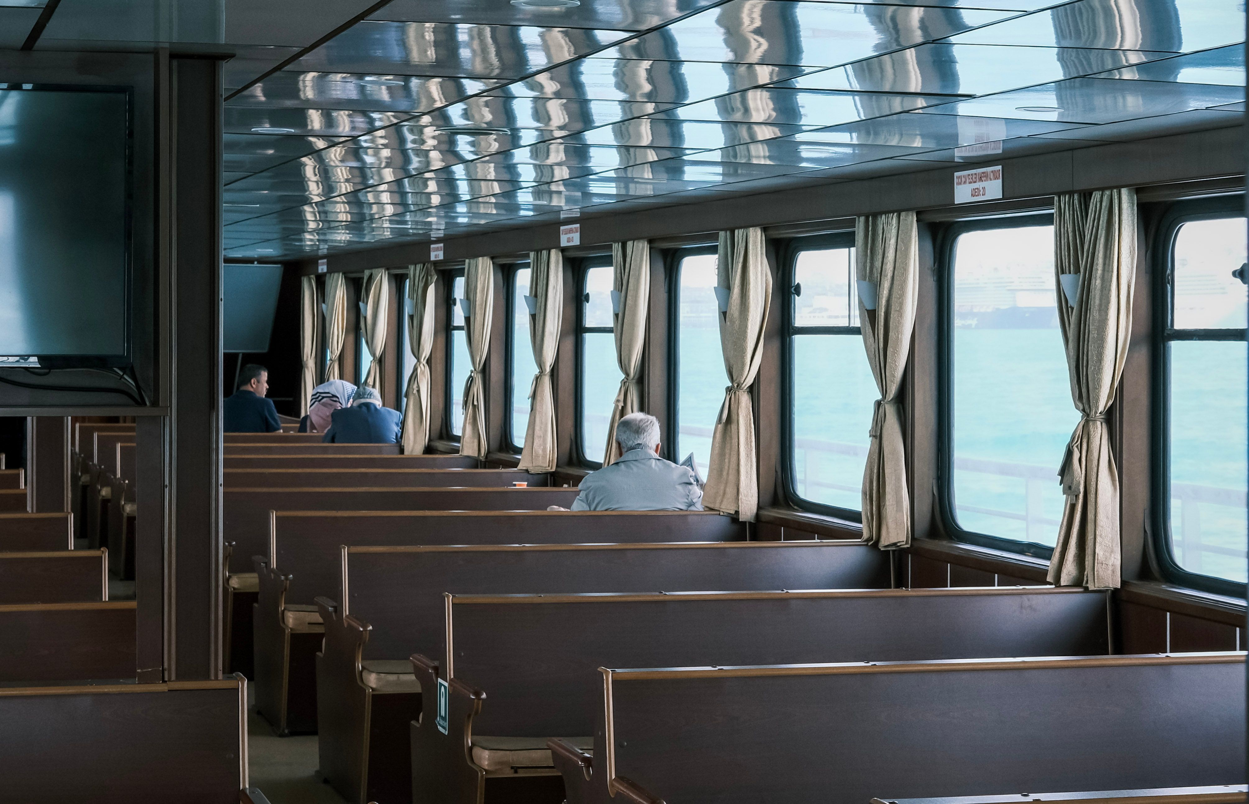 A quiet ferry interior with wooden seating and a few passengers. Daylight streaming through the windows.
