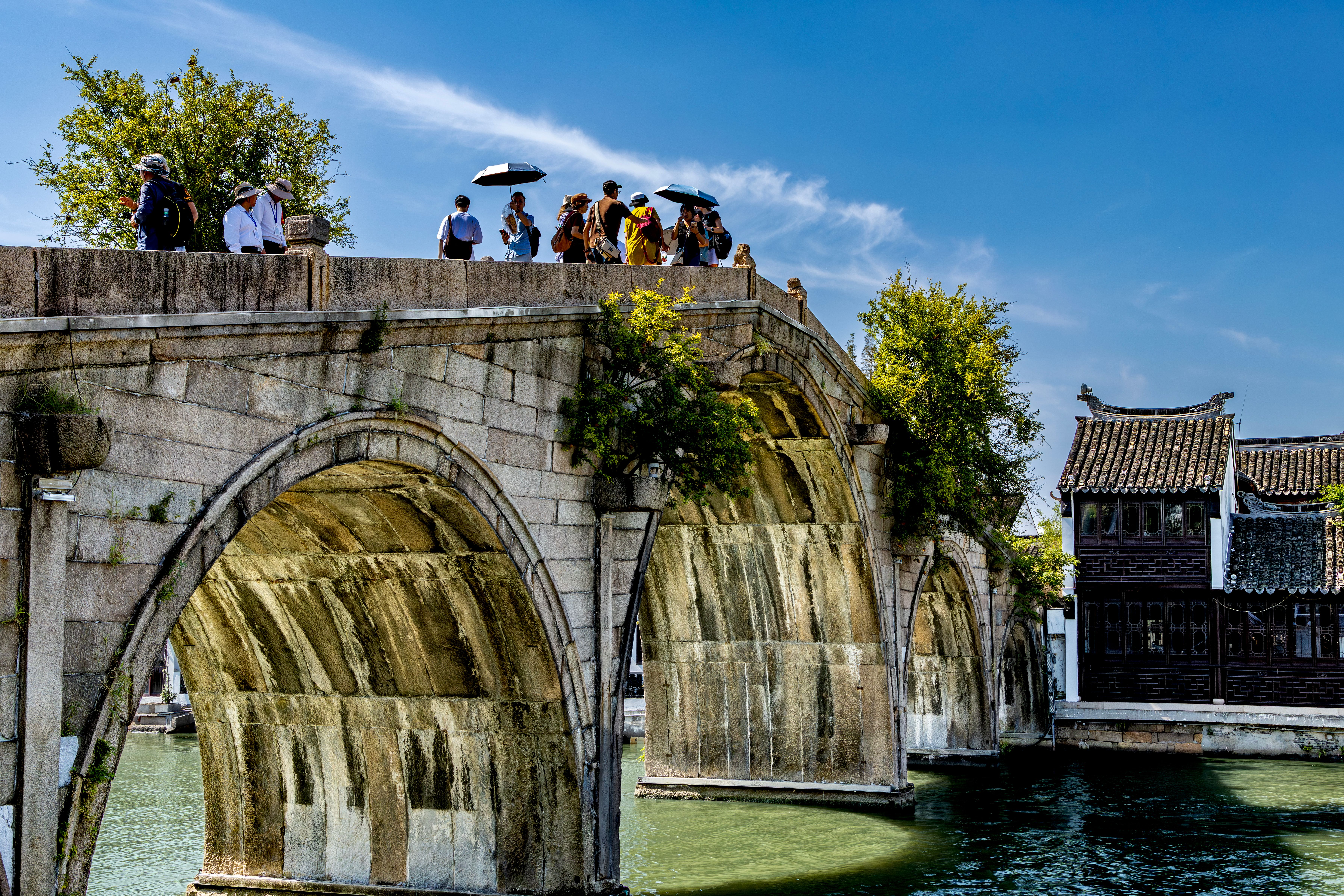Tourists on an ancient stone arch bridge in a traditional Asian water town under a clear blue sky.