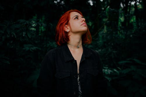 Pensive young female with red hair in casual clothes standing in lush green jungle and looking up