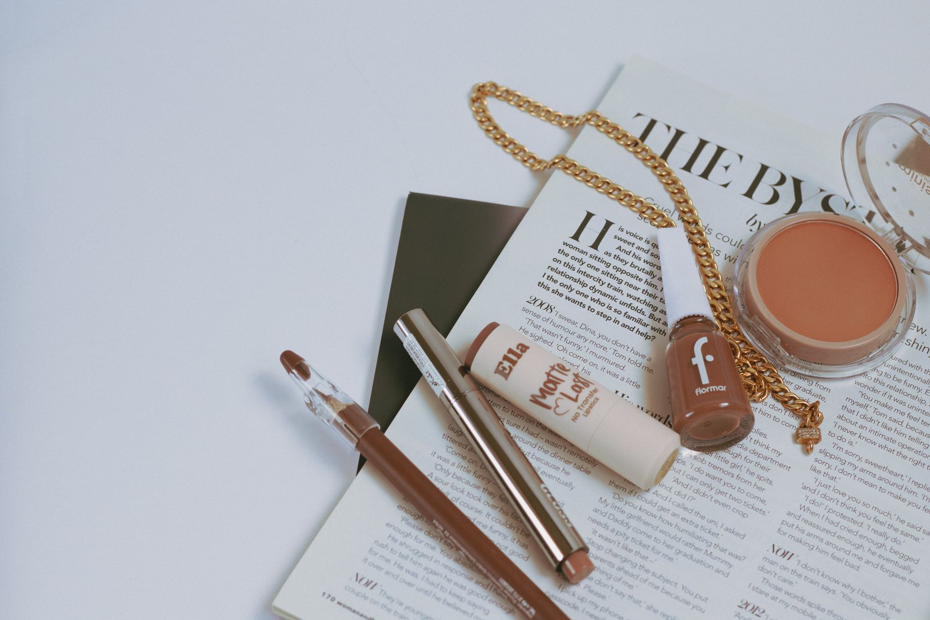 Flat lay of makeup products on a magazine layout with gold chain accessory.
