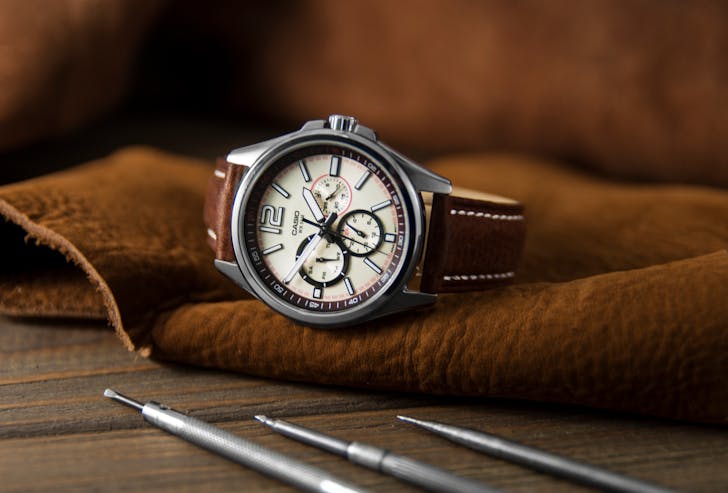 Stylish round wristwatch with chronograph and brown strap placed on leather material near repair tools in modern workshop