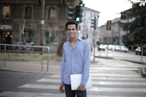 Positive ethnic guy in shirt holding laptop and smiling at camera while standing on city pedestrian crosswalk in city center