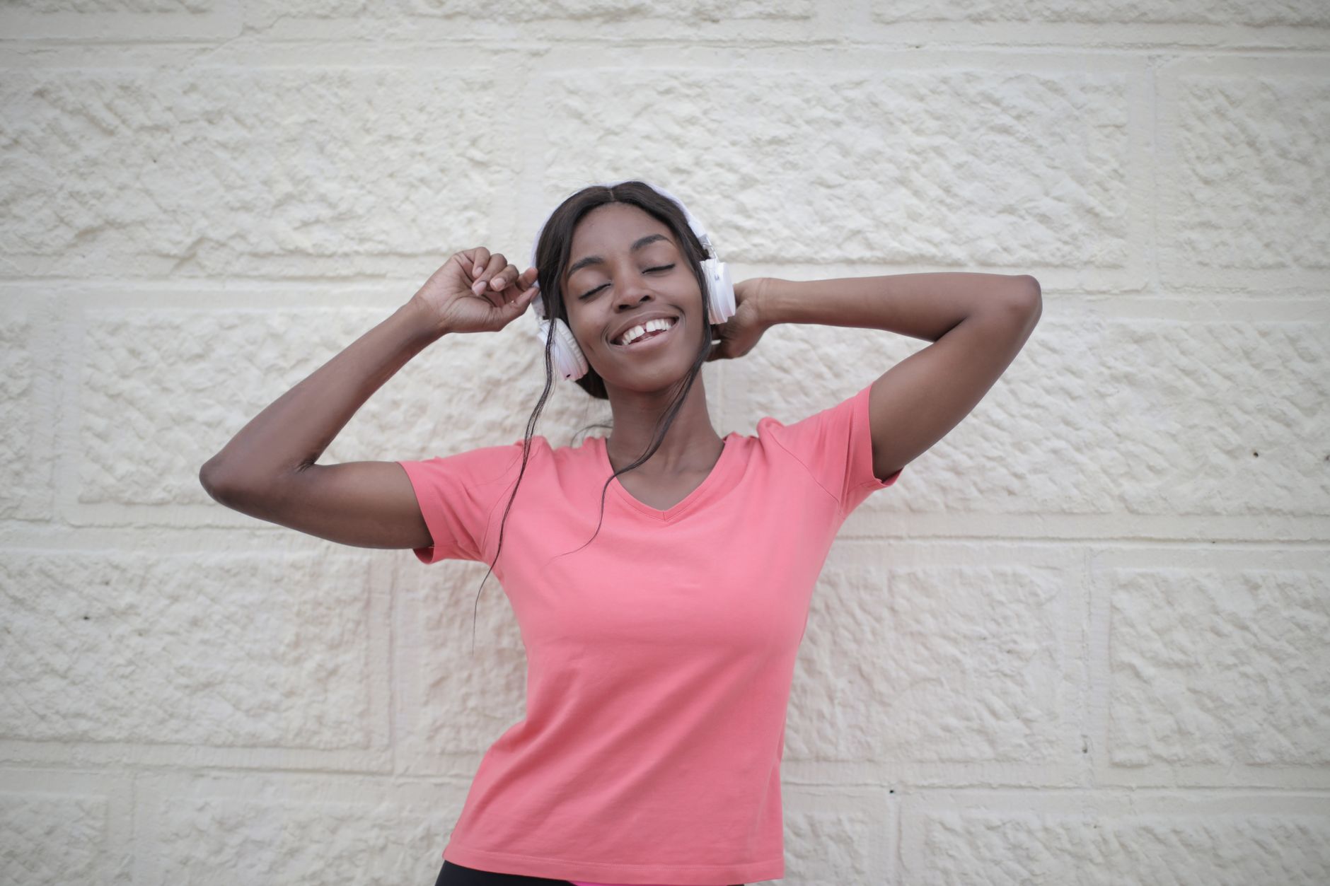 Happy African American female in pink t shirt and headphones enjoying music and dancing with eyes closed against white wall