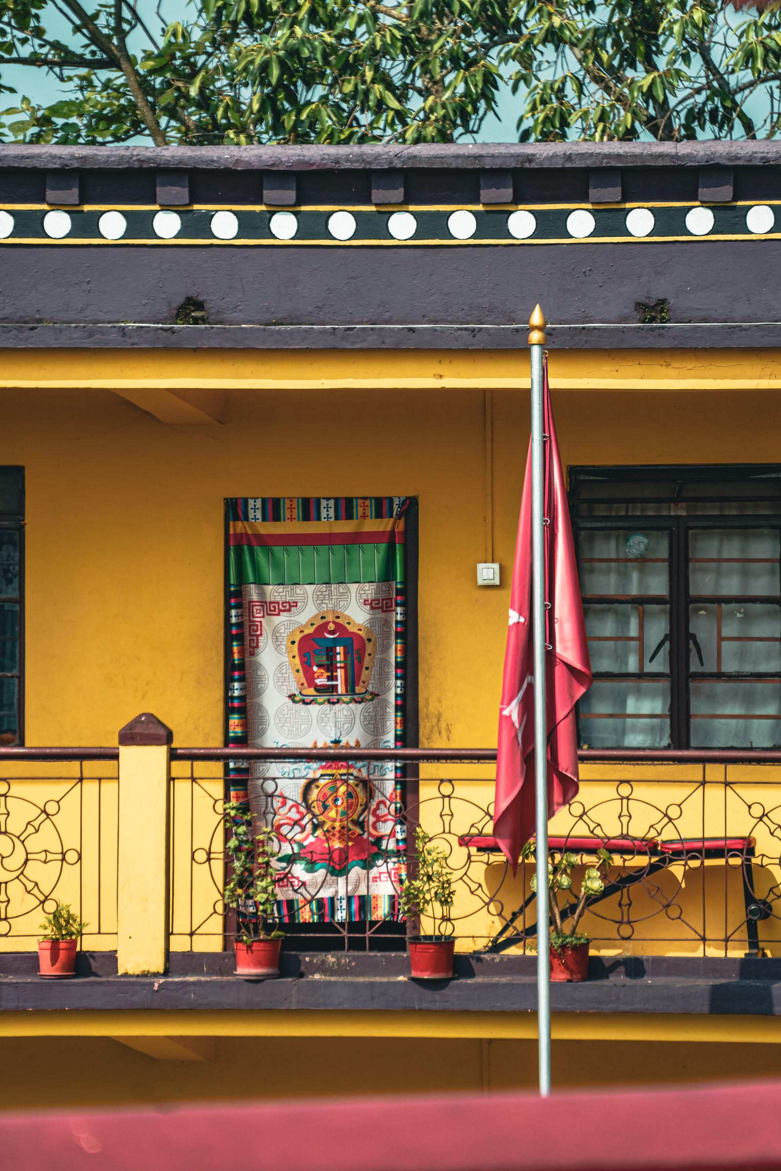 A yellow Tibetan Buddhist balcony in Kalimpong hung with a painted door curtain, dharma wheel railings and a maroon flag