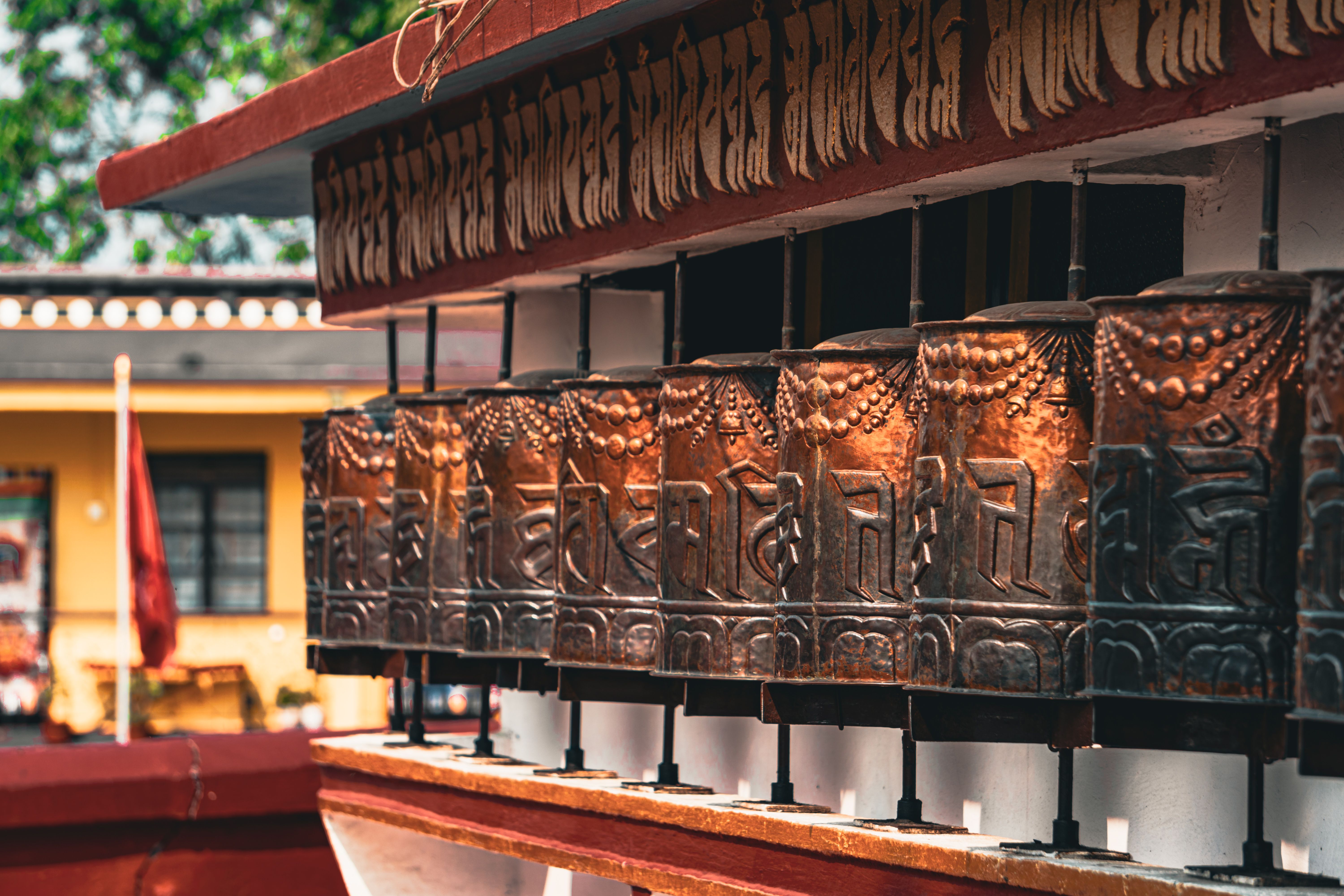 Carved copper prayer wheels in a row under a red eave lettered in Tibetan script, at a monastery in Kalimpong