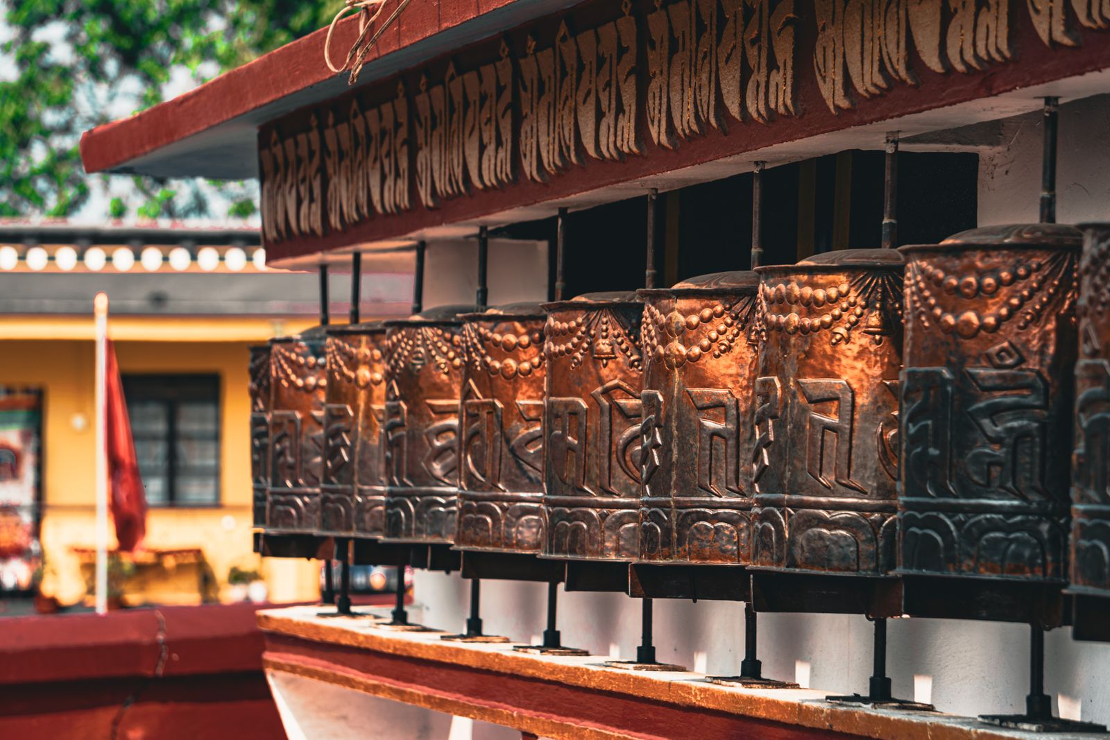 A row of carved copper prayer wheels under a red eave lettered in Tibetan, at a monastery in Kalimpong