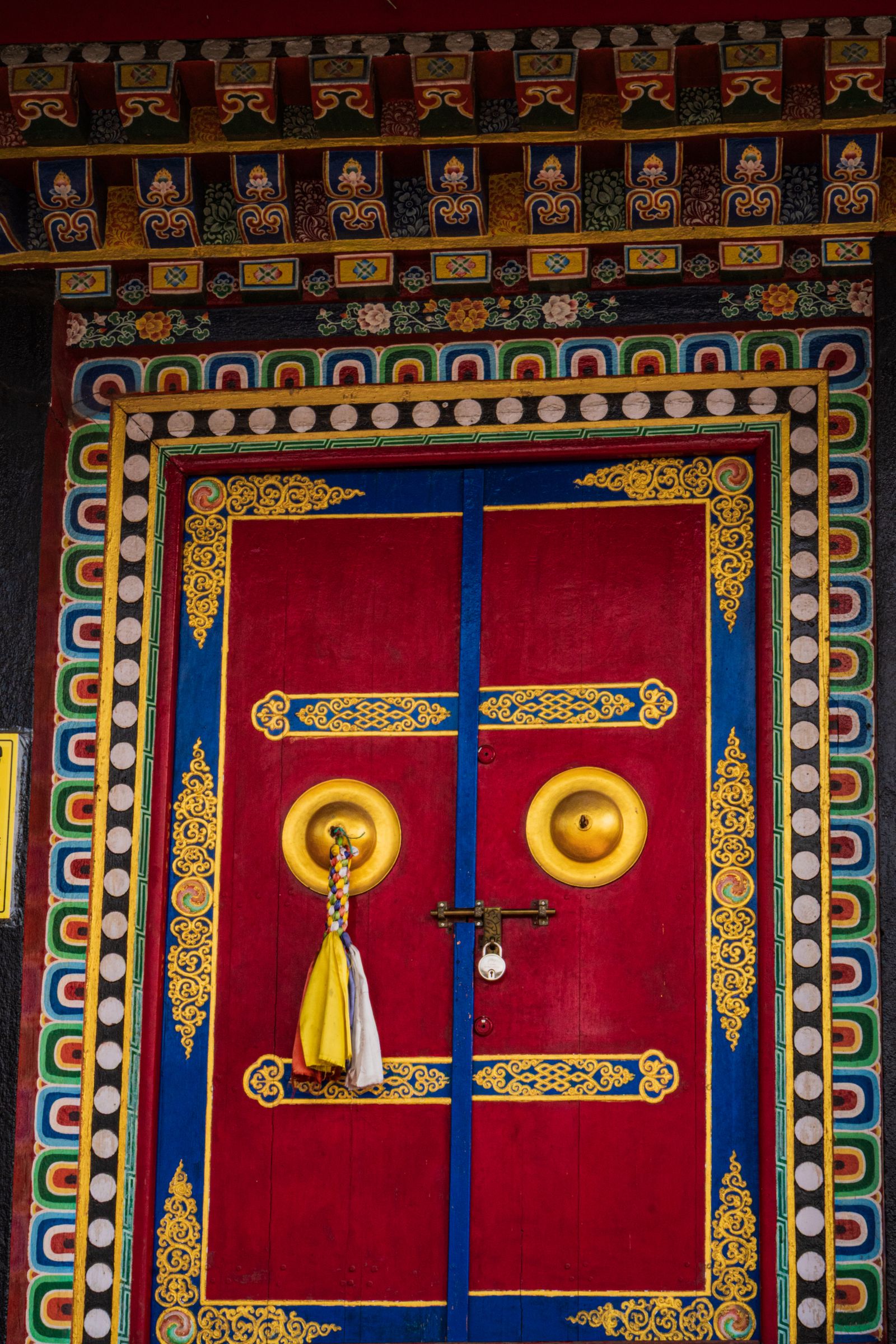 A red and gold painted monastery door with brass rings and a silk tassel, framed by bright carved borders in Kalimpong