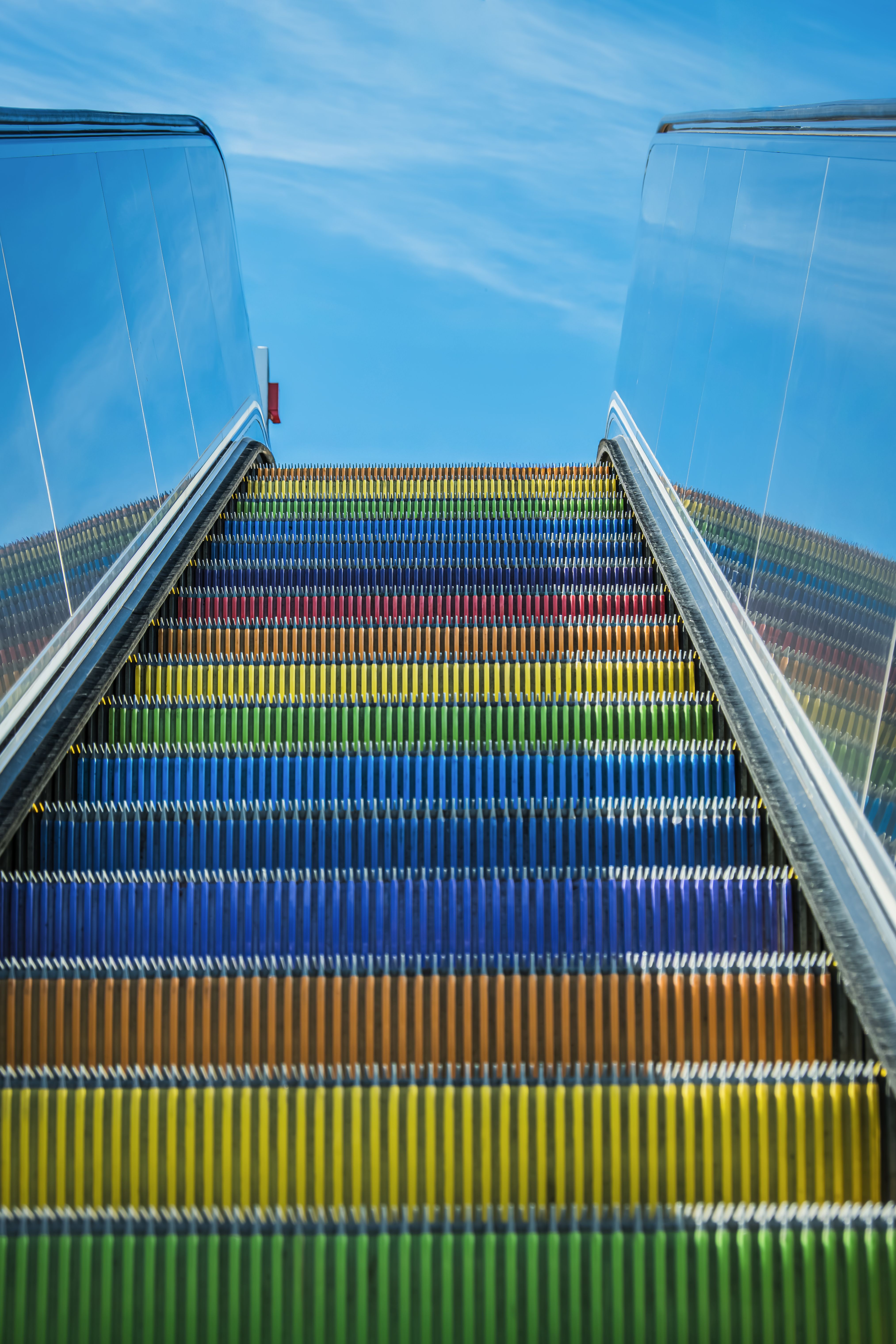 Vibrant rainbow-colored escalator steps lead upwards against a bright blue sky.