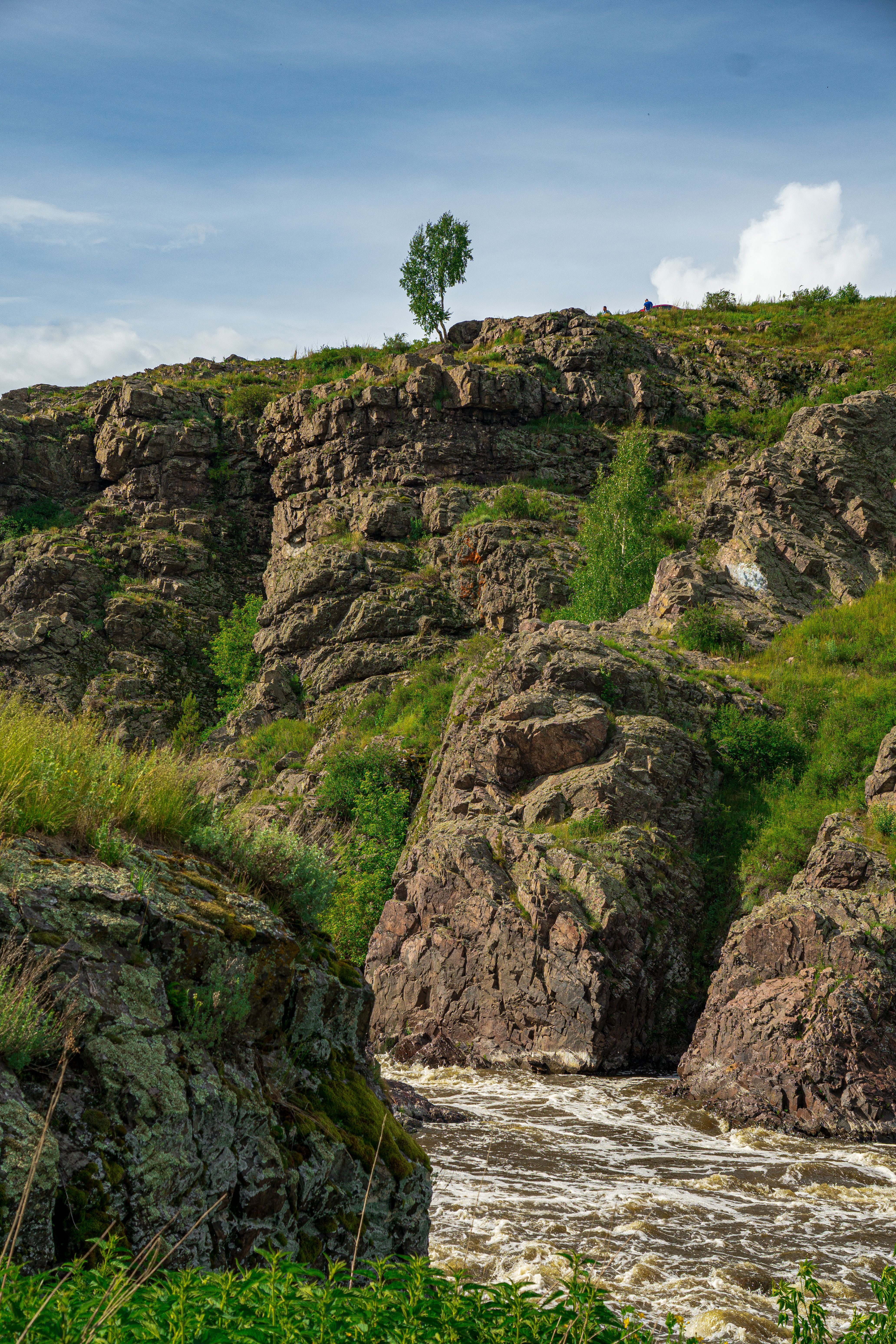 A picturesque rocky hillside with lush greenery and river cascading below under a clear blue sky.