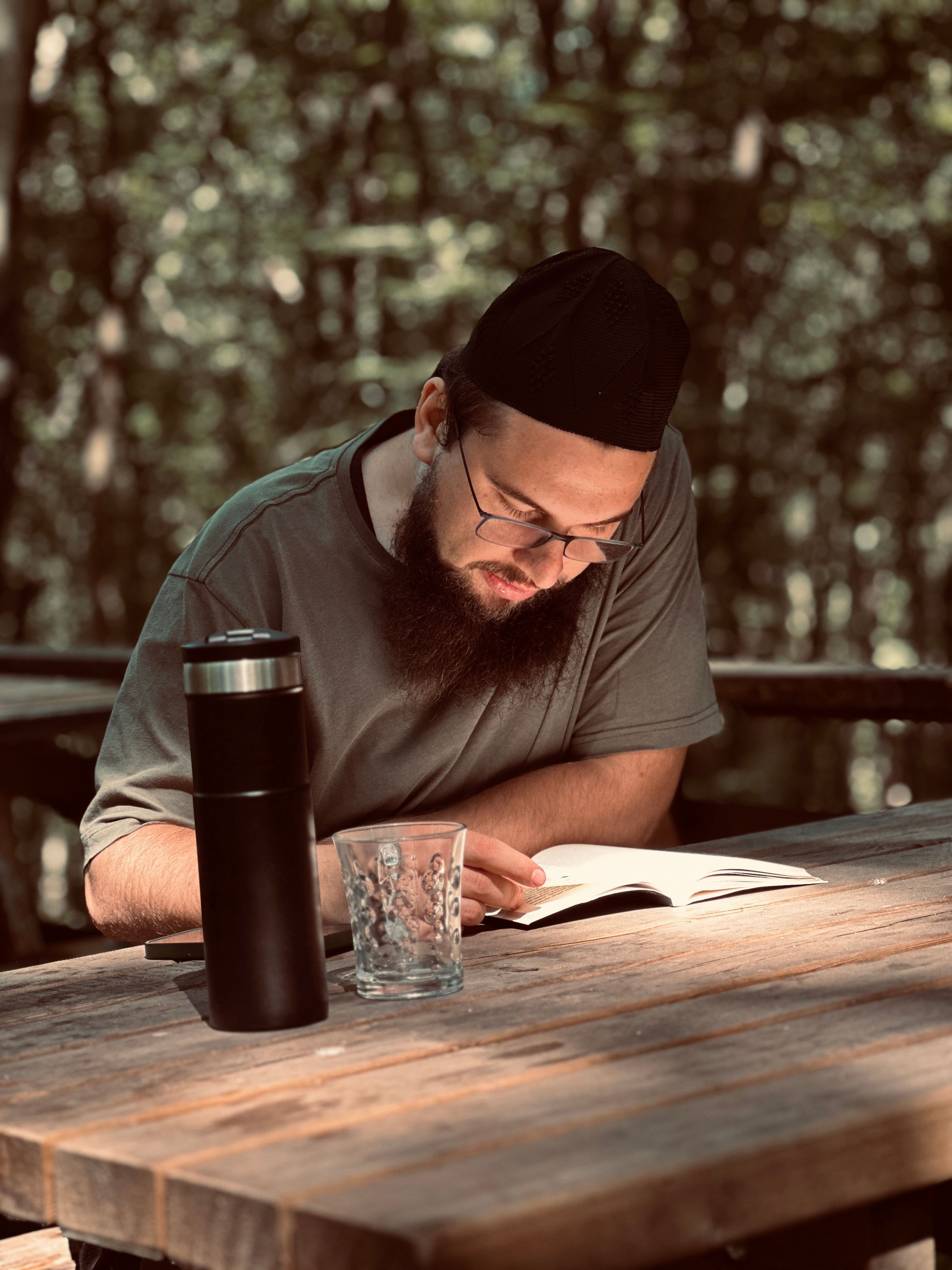 Bearded man reading outdoors in a serene forest setting with a thermos and glass on the table.