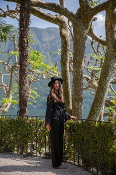 Woman in a stylish black outfit standing in a lush garden with scenic mountain and lake view.