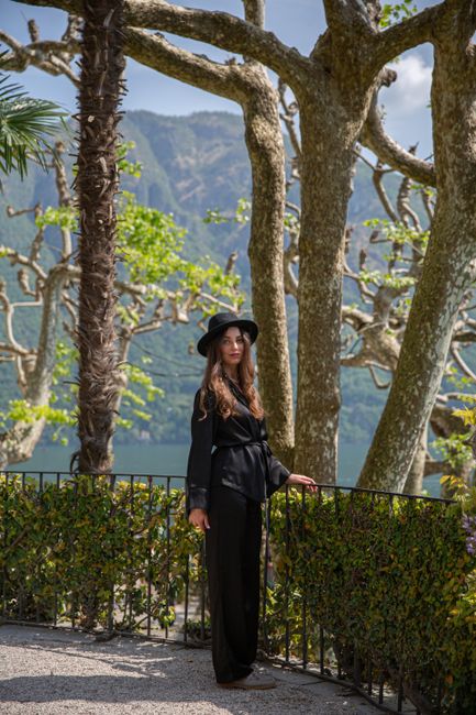 Woman in a stylish black outfit standing in a lush garden with scenic mountain and lake view.