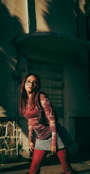 A stylish young woman wearing glasses poses against a building in an urban setting during the day.