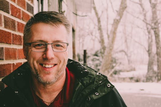 一位戴着眼镜、留着胡须、面带微笑的男子，身穿冬衣，站在白雪皑皑的户外砖墙旁