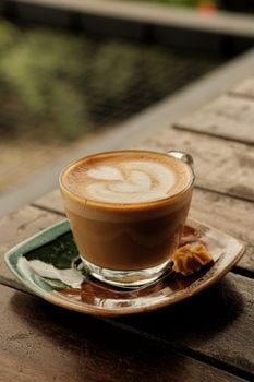 A creamy latte with intricate art in a clear glass cup on a rustic wooden table.