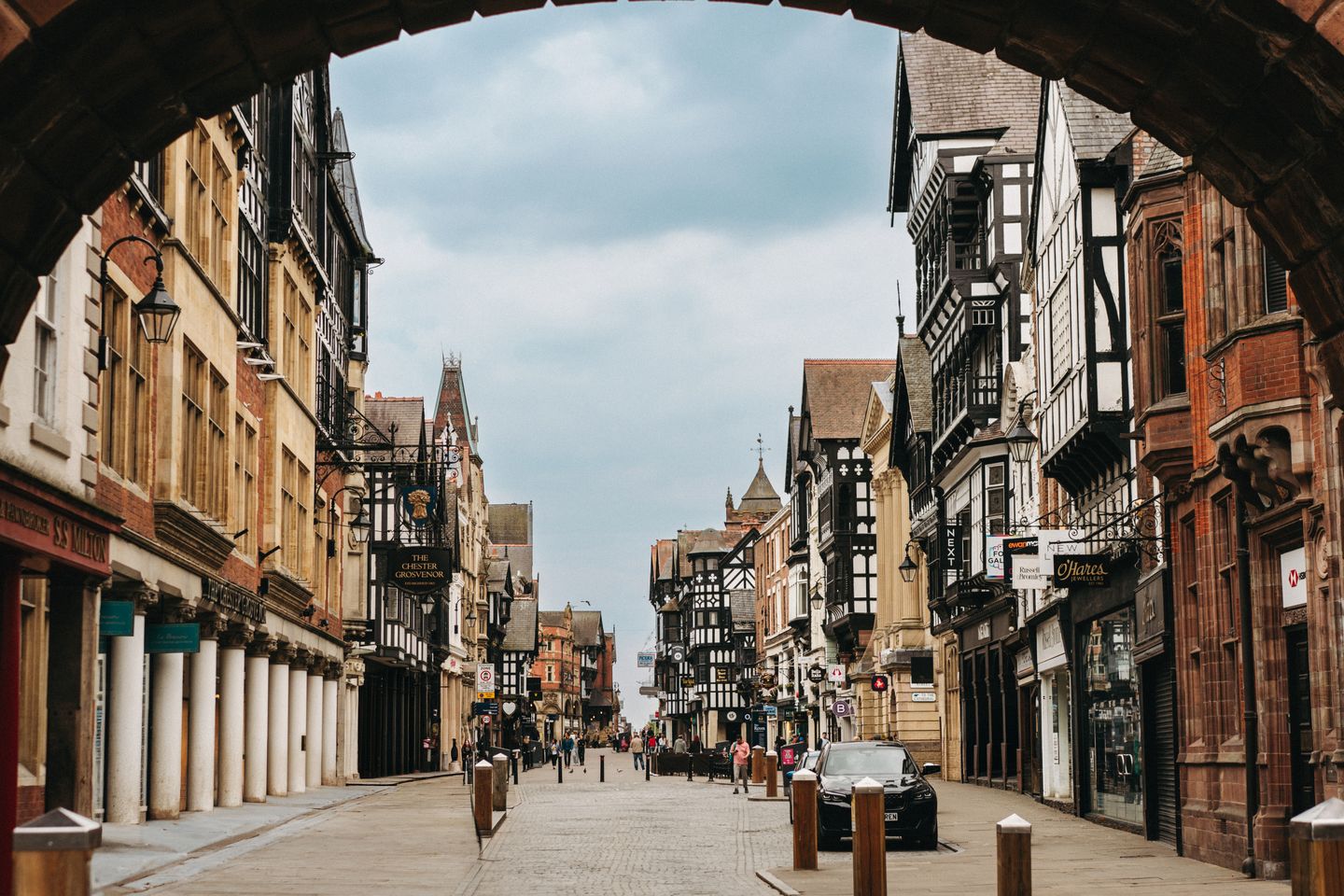 Chester's half-timbered high street through a stone arch, at an hour when people still go into shops to try things on.
