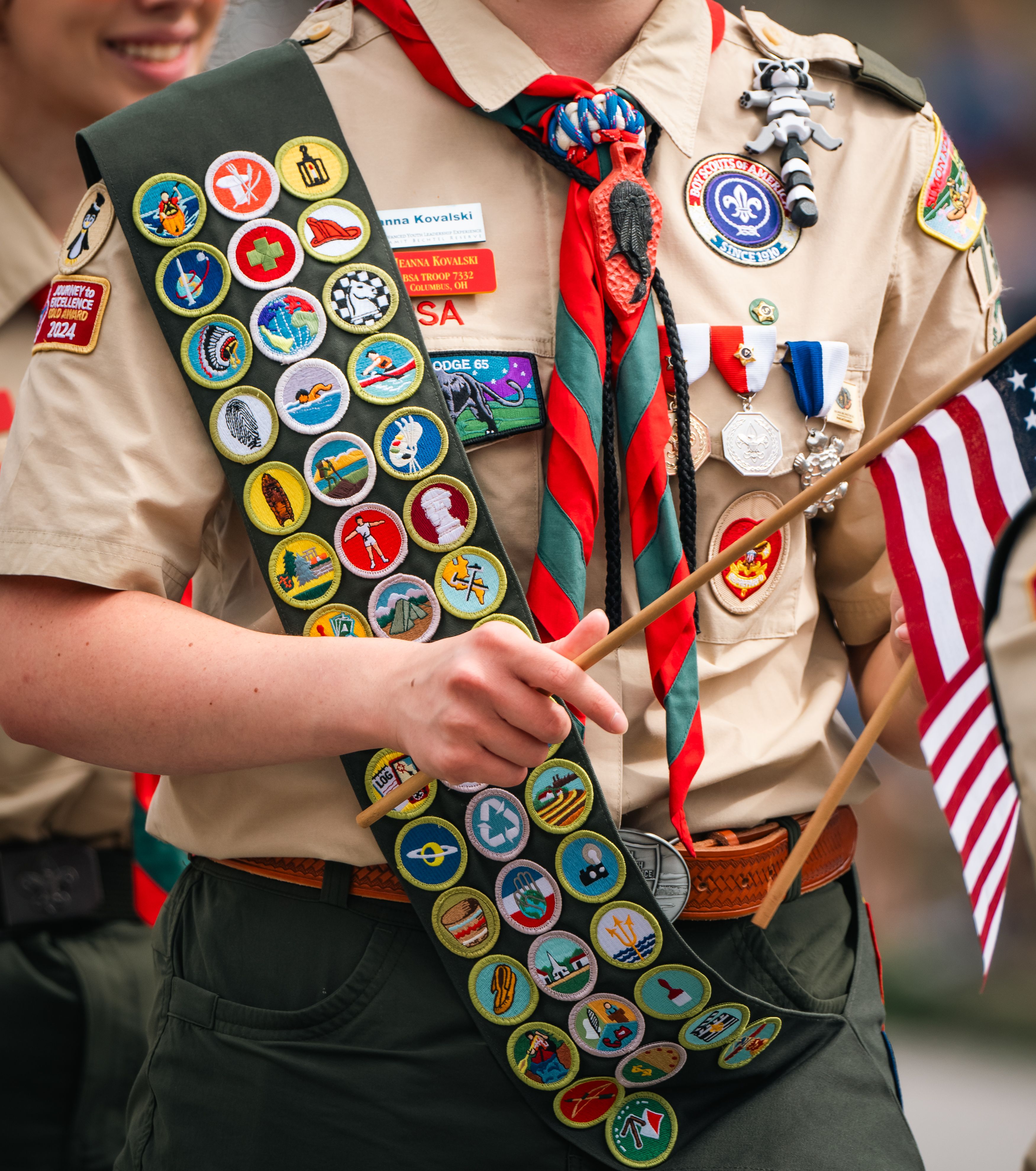 Close-up of a Boy Scout in uniform with a merit badge sash, holding a small American flag.