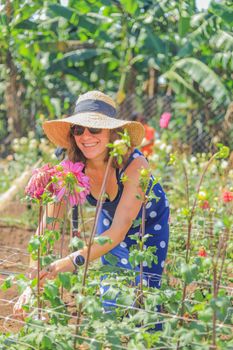 Smiling woman in sun hat tending flowers in a bright garden, enjoying a sunny day outdoors.