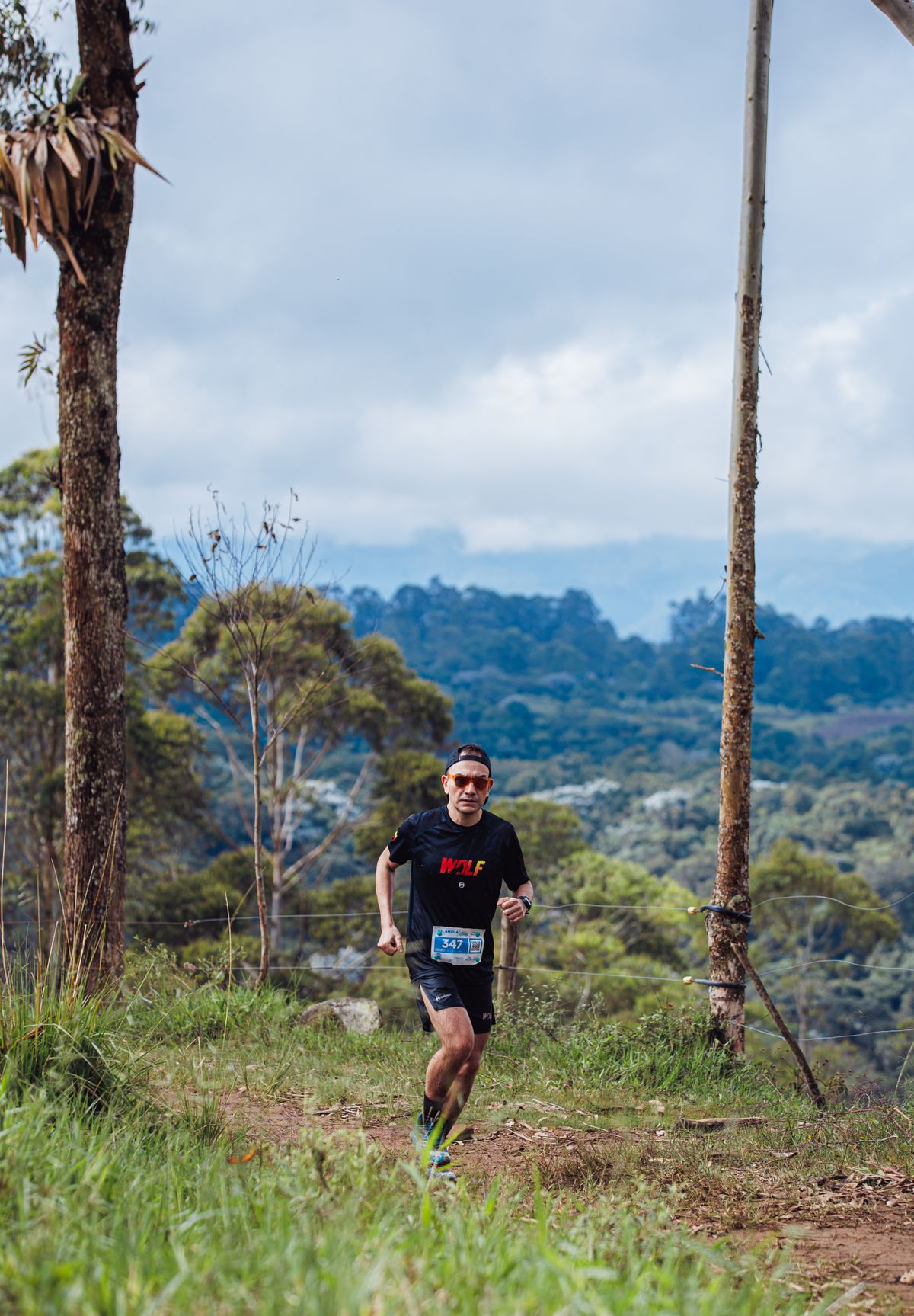 A lone trail runner on a mountain path surrounded by green forest under a cloudy sky