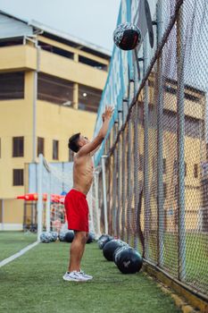 Shirtless man in red shorts doing wall ball workout on a field.