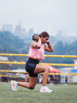 Woman performing a lunge with a dumbbell outdoors on a field against a cityscape.