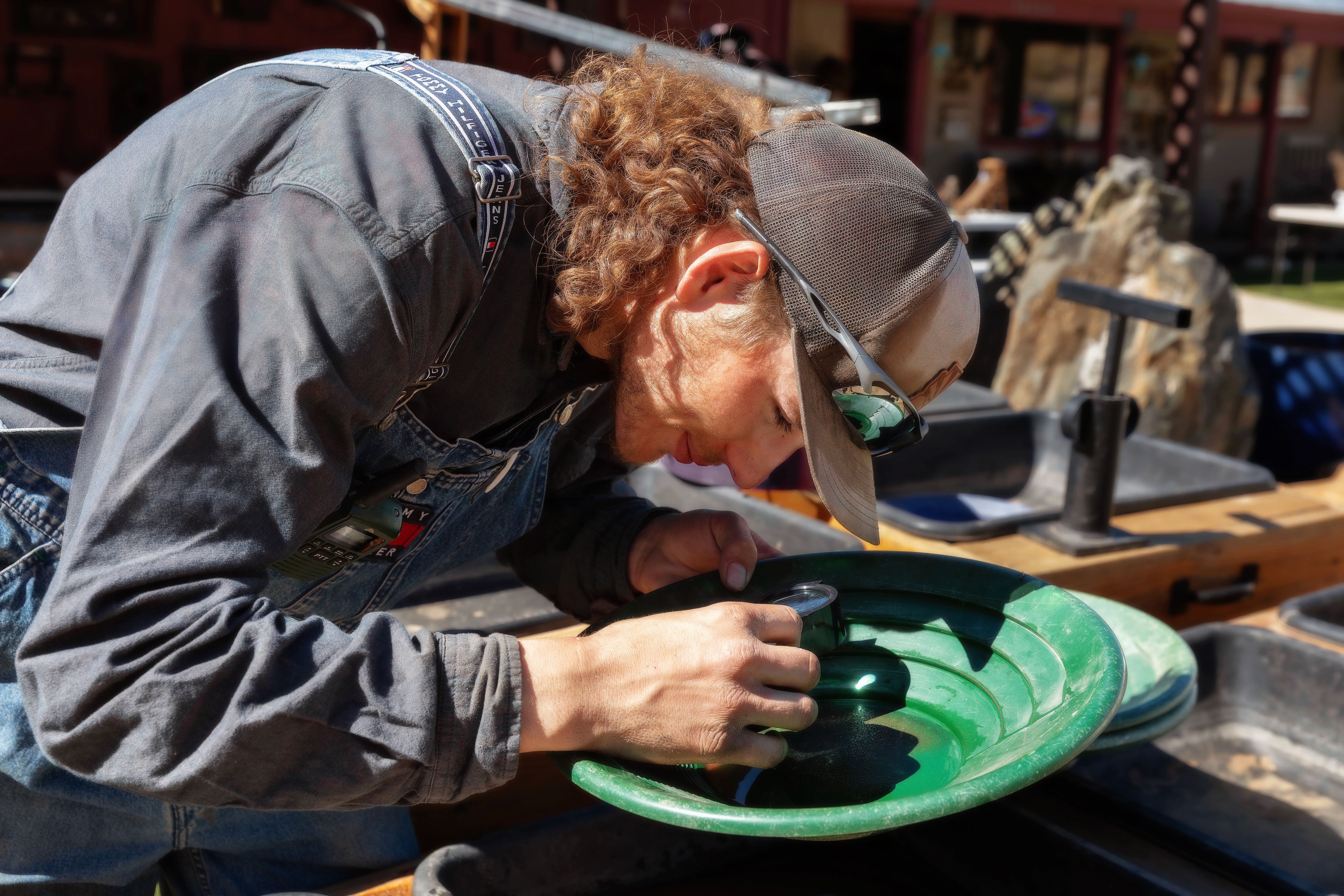 A man in overalls uses a gold pan under sunlight, focusing intently outdoors.