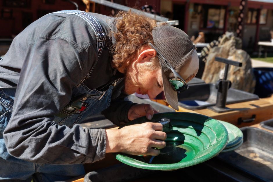 A man in overalls uses a gold pan under sunlight, focusing intently outdoors.