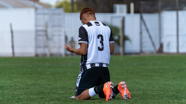 A young male soccer player kneels in prayer or reflection on the grass field.
