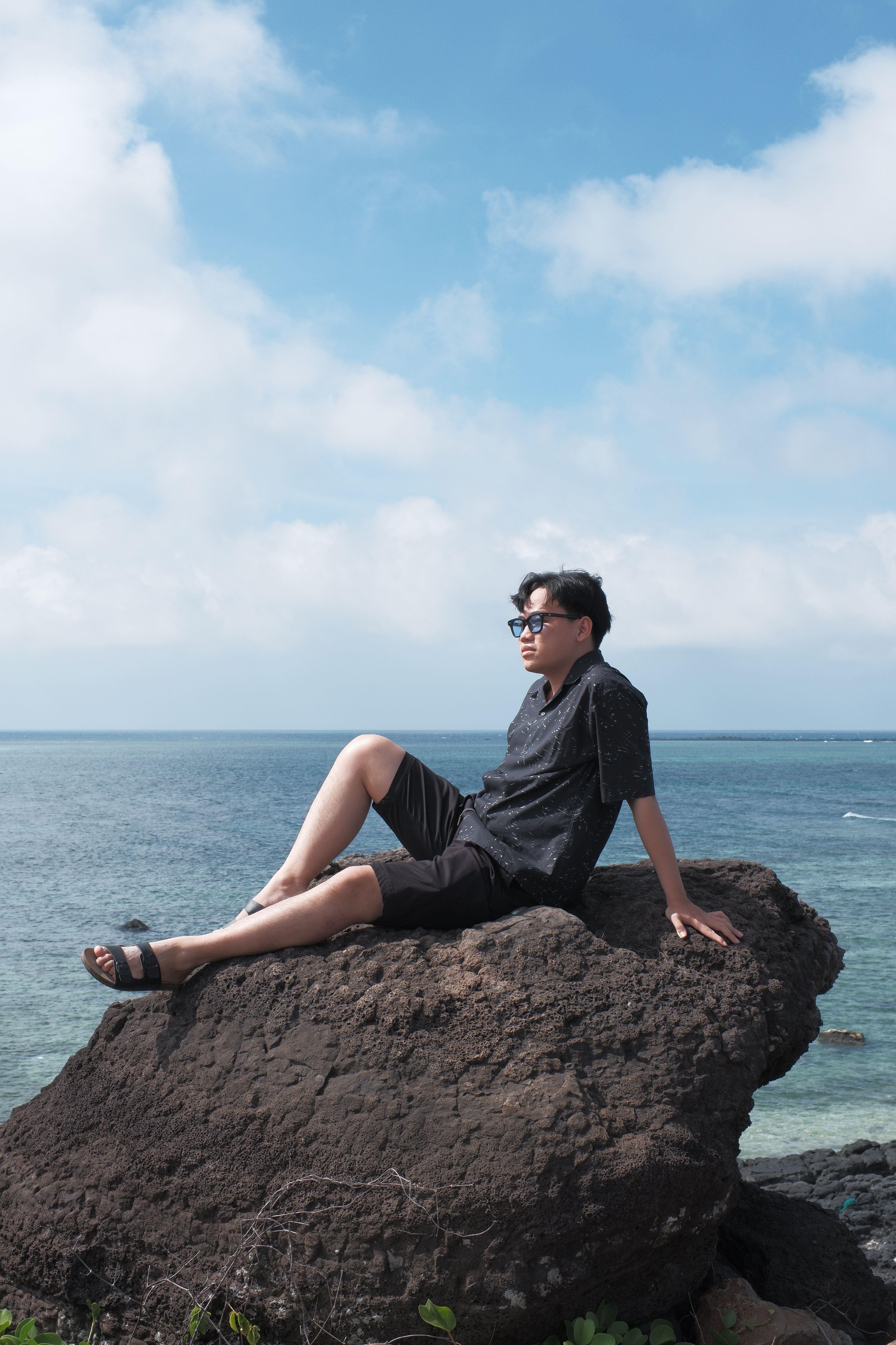 A man enjoys a peaceful moment sitting on a large rock by the ocean, under a clear blue sky.