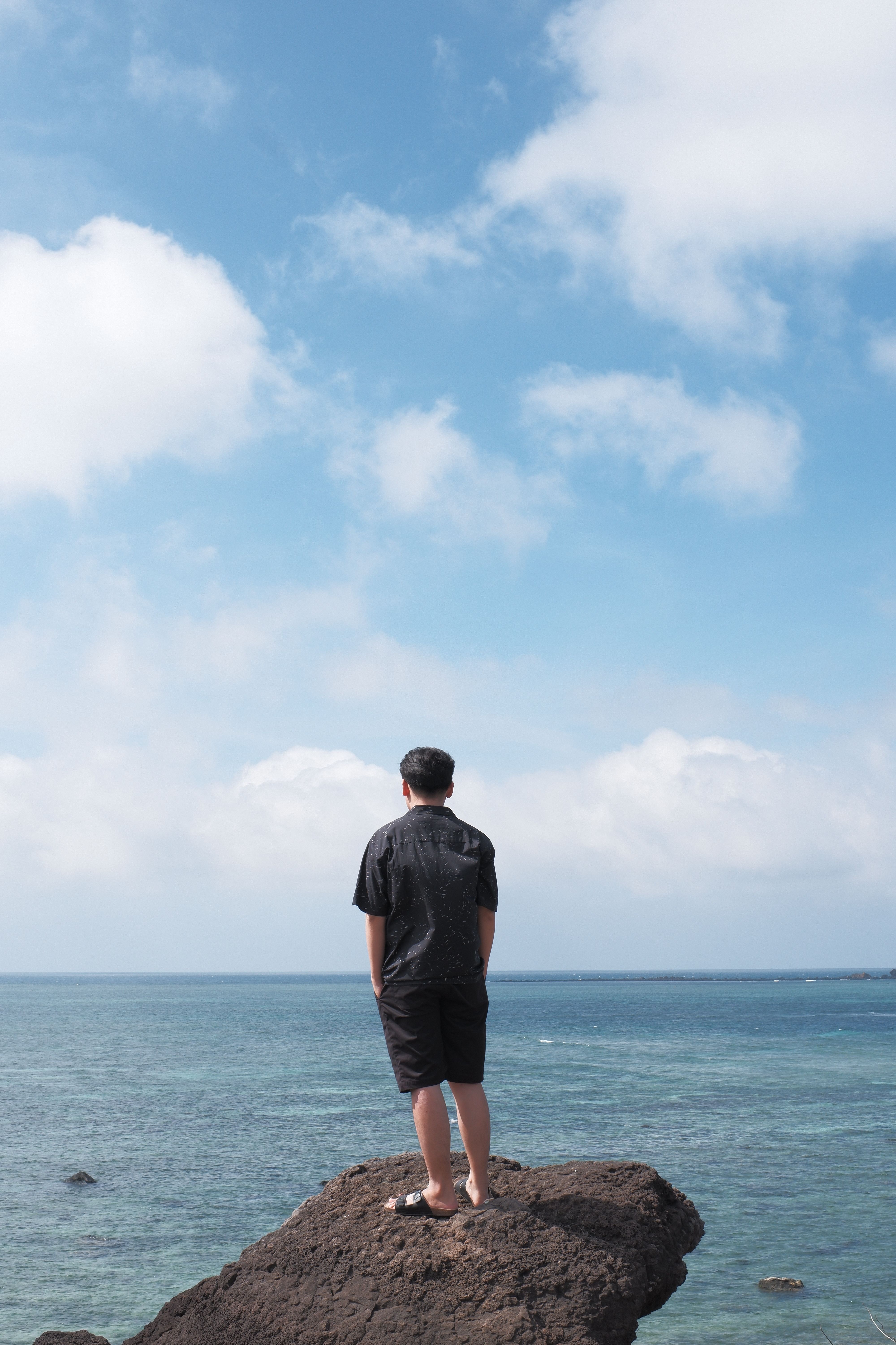 A man stands on a rocky ledge gazing at the calm ocean under a clear blue sky.