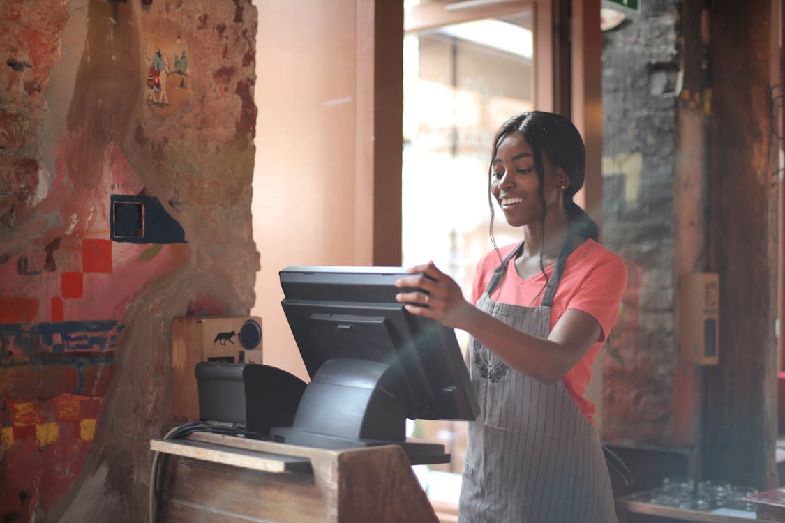 Free Cheerful American African waitress in apron working on counter monitor while registering order at cozy cafe Stock Photo