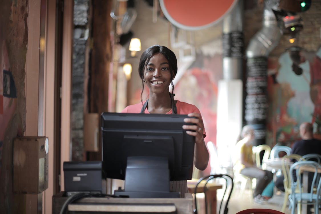 Free Positive young woman in uniform smiling while standing at counter desk in cafe Stock Photo