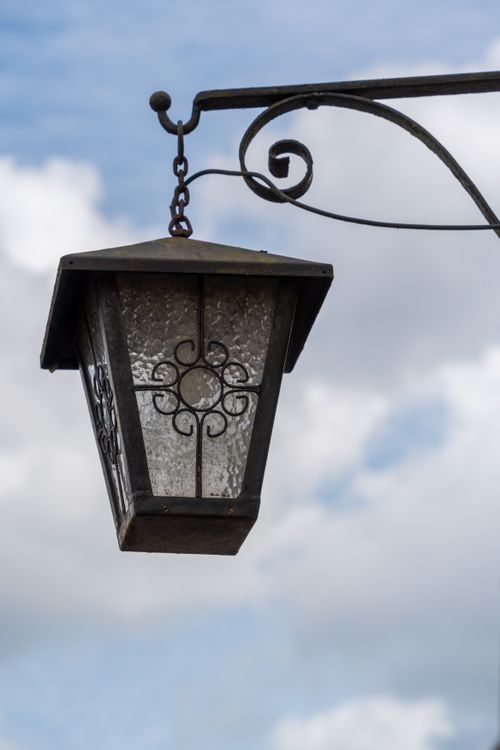Free Close-up of a vintage wrought iron lantern with decorative glass against a blue sky with clouds. Stock Photo