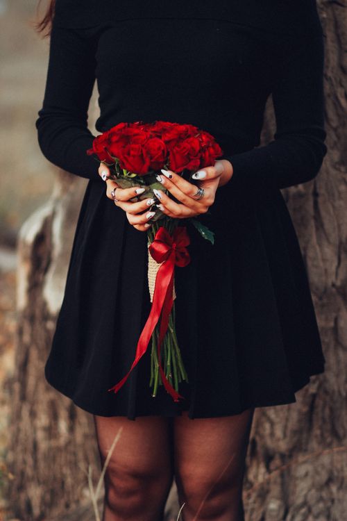Free A woman in black dress holding a vibrant red rose bouquet with a ribbon, set against a natural outdoor backdrop. Stock Photo