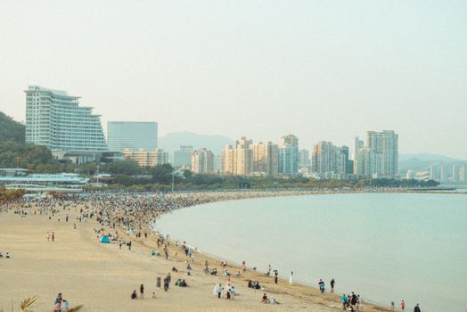 A bustling beach scene with a city skyline under a clear sky.