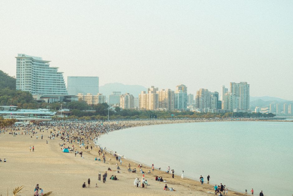 A bustling beach scene with a city skyline under a clear sky.