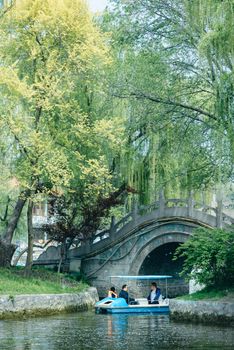 Two adults enjoy a peaceful boat ride under a stone bridge surrounded by lush spring foliage.